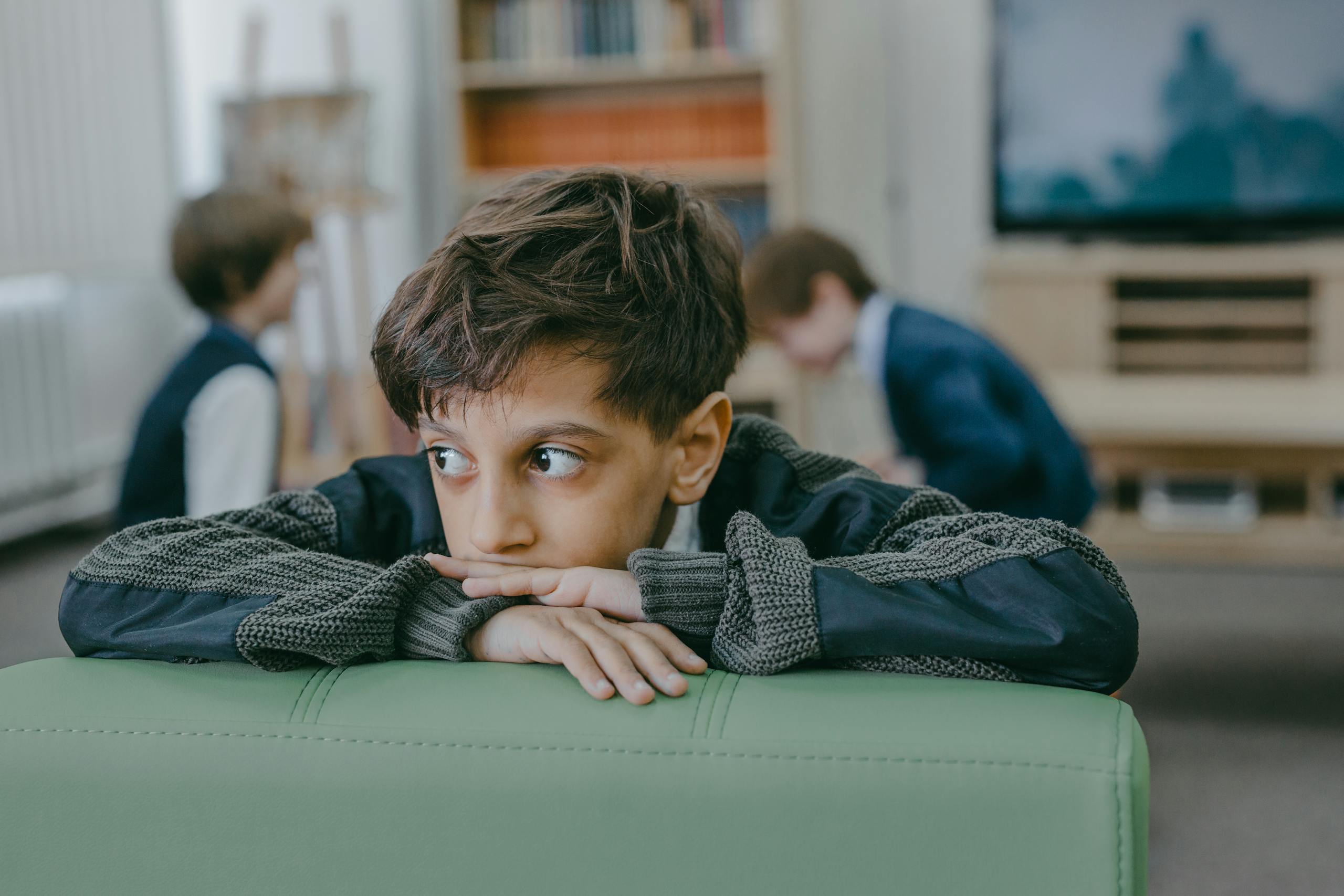 A young boy leans on a green chair with a thoughtful expression, indoors.